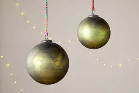 Two Oko Giant Baubles - Matt Green by nkuku hang against a beige background, accented with faint string lights. These festive Christmas decorations feature gold and brown details and are each suspended by a colorful string.