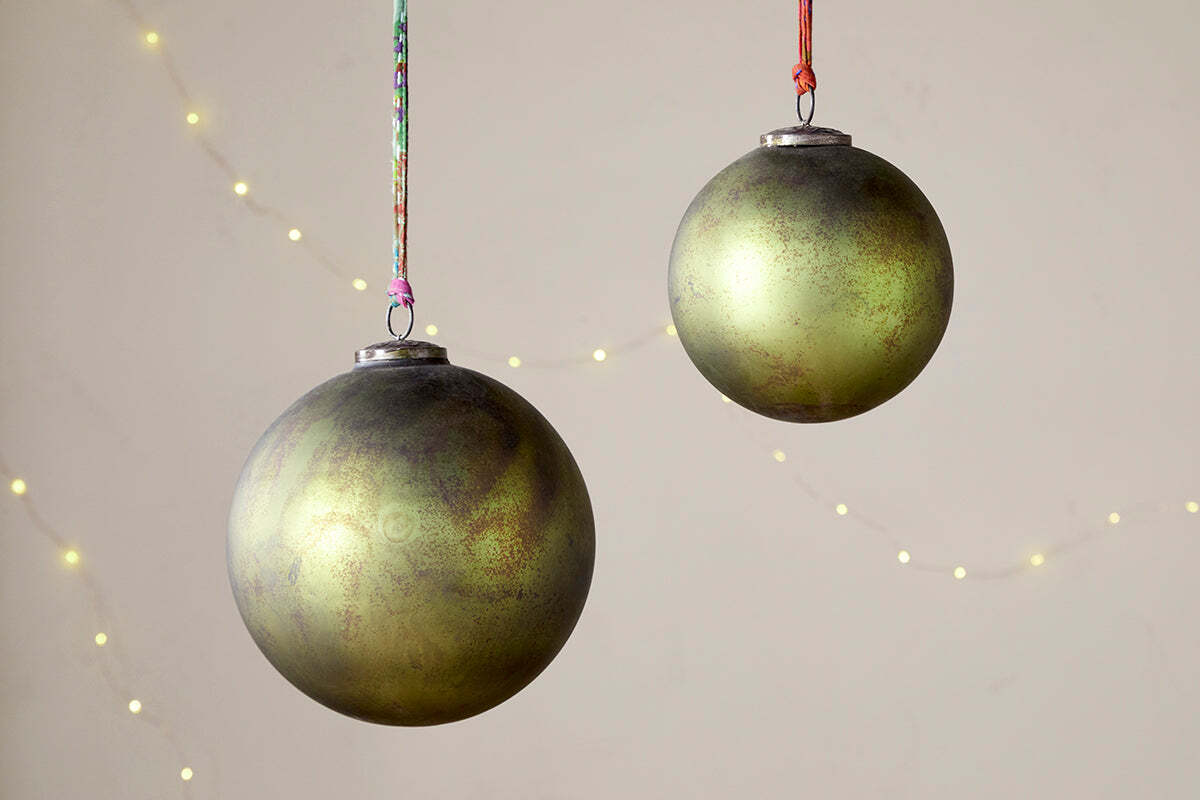 Two Oko Giant Baubles - Matt Green by nkuku hang against a beige background, accented with faint string lights. These festive Christmas decorations feature gold and brown details and are each suspended by a colorful string.