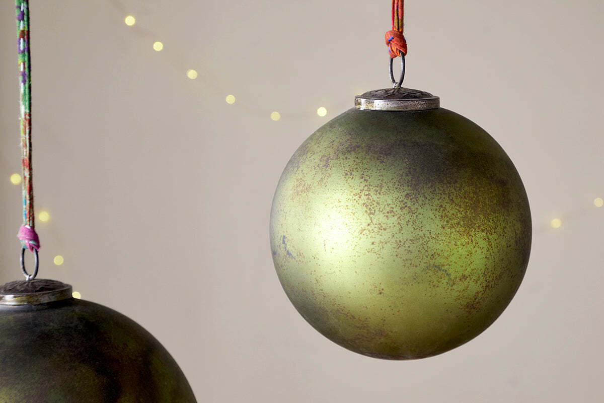 Two large Oko Giant Bauble - Matt Green ornaments by nkuku hang from colorful strings against a neutral background with soft yellow lights. Their slightly tarnished, vintage look adds rustic charm to this festive scene.