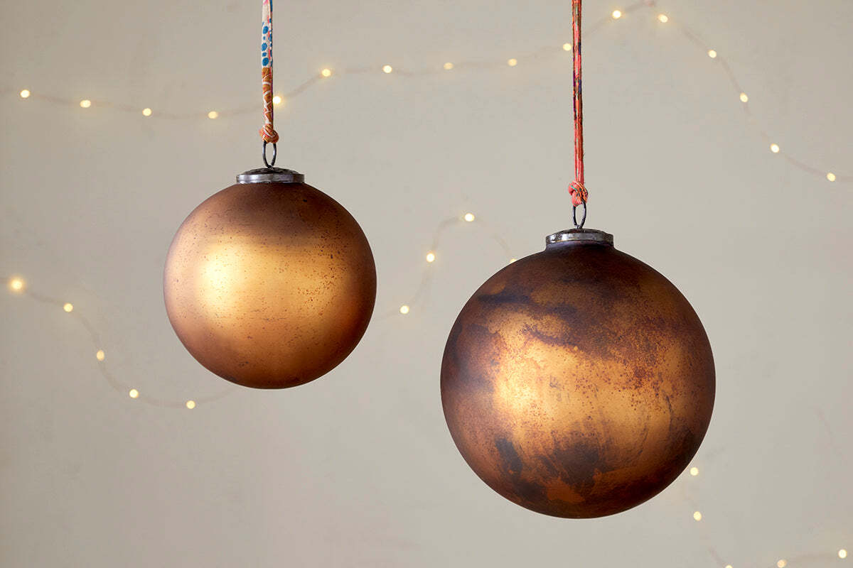 The nkuku Oko Giant Bauble in Matt Gold hangs from a colorful string against a neutral backdrop with warm white fairy lights, creating a cozy, festive holiday atmosphere.