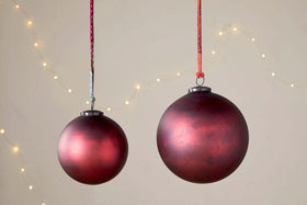 Two nkuku Oko Giant Baubles in Matt Bordeaux hang from colorful strings against a neutral background. One bauble is larger, and small, blurred string lights create a soft, festive display behind the Christmas decorations.
