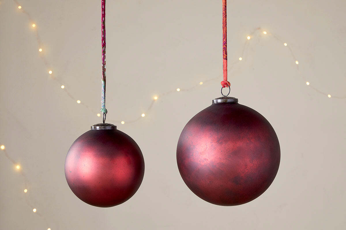 Two nkuku Oko Giant Baubles in Matt Bordeaux hang from colorful strings against a neutral background. One bauble is larger, and small, blurred string lights create a soft, festive display behind the Christmas decorations.