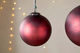 A close-up of the nkuku Oko Giant Bauble in Matt Bordeaux hangs against a neutral backdrop with softly blurred yellow fairy lights, while a second bauble is partly visible on the image’s right edge for a festive touch.