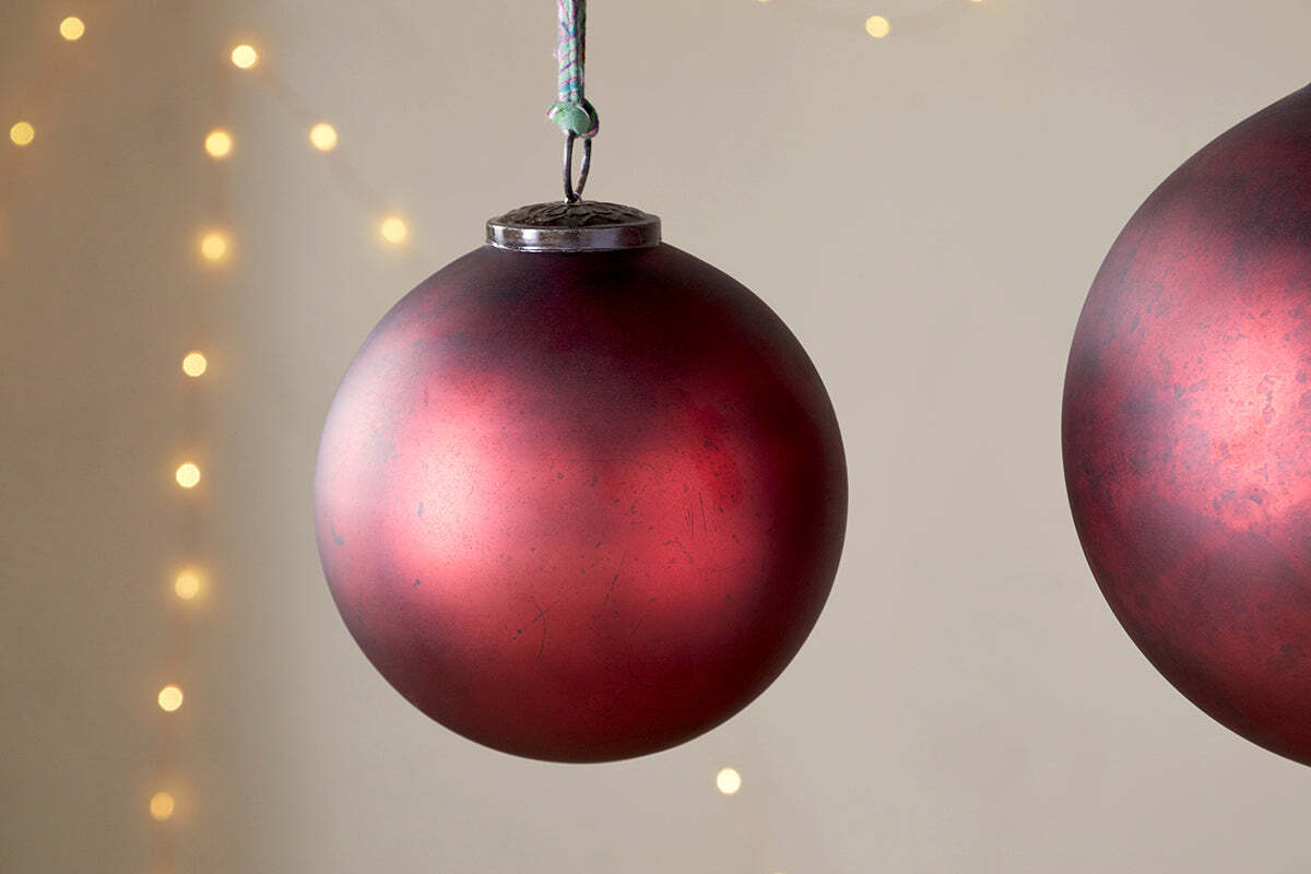 A close-up of the nkuku Oko Giant Bauble in Matt Bordeaux hangs against a neutral backdrop with softly blurred yellow fairy lights, while a second bauble is partly visible on the image’s right edge for a festive touch.