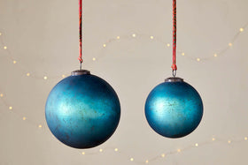 The nkuku Oko Giant Bauble in Matt Teal hangs alongside a smaller ornament on red strings, set against a neutral backdrop with soft string lights. Their metallic finish and weathered look create an inviting festive atmosphere.