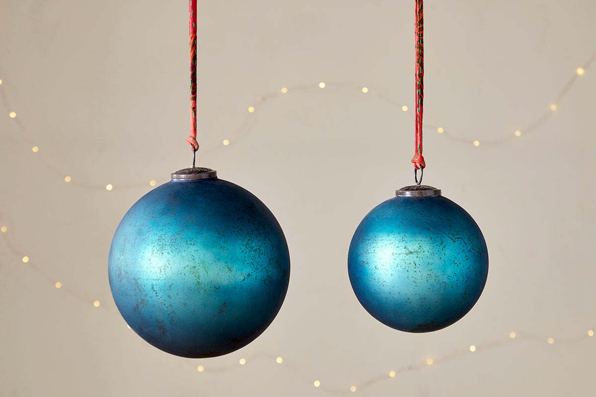 The nkuku Oko Giant Bauble in Matt Teal hangs alongside a smaller ornament on red strings, set against a neutral backdrop with soft string lights. Their metallic finish and weathered look create an inviting festive atmosphere.