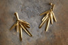 The nkuku Omuti Earrings - Gold feature multiple textured, elongated leaf-like bars with a radiant gold finish, offering chic, contemporary style—perfect party-ready accessories shown on a rough stone surface.