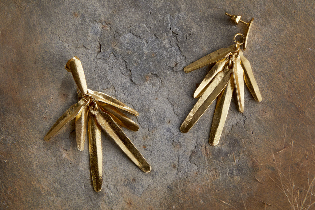 The nkuku Omuti Earrings - Gold feature multiple textured, elongated leaf-like bars with a radiant gold finish, offering chic, contemporary style—perfect party-ready accessories shown on a rough stone surface.