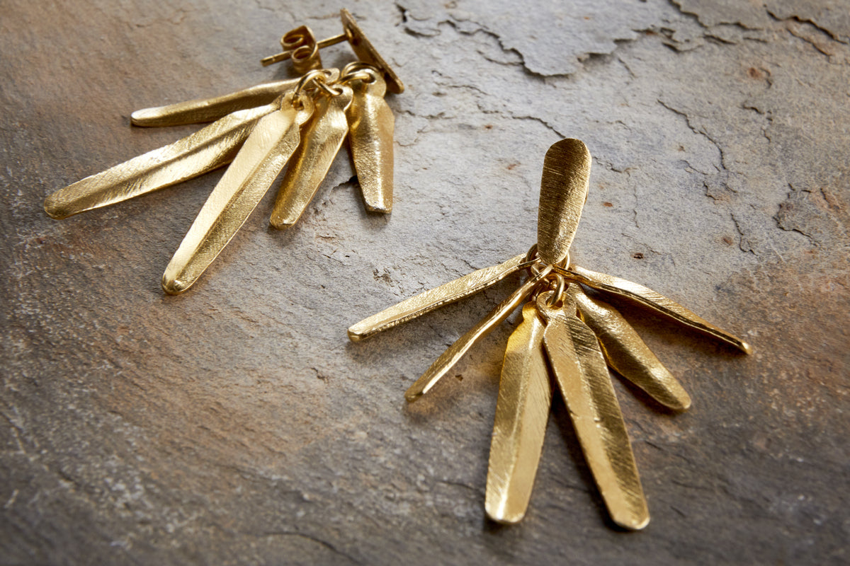The nkuku Omuti Earrings - Gold showcase multiple elongated, textured metal pieces with a gold-coloured finish, styled on a stone surface—ideal for lovers of contemporary jewellery.