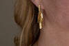 Close-up of a person wearing the Omuti Earrings - Gold by nkuku, featuring multi-layered, leaf-shaped design with a gold finish. The party-ready earrings hang elegantly, with light brown hair and skin in the background.