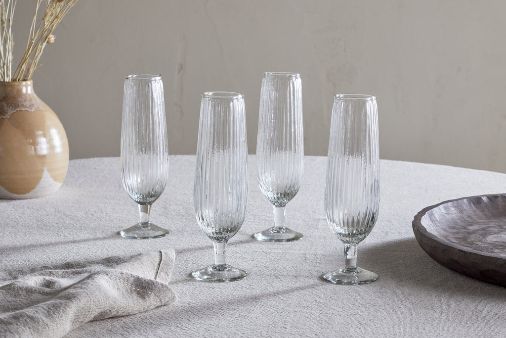 A set of four Otuba Champagne Flutes in clear, by nkuku, are arranged on a light tablecloth, with a ceramic vase of dried plants and a wooden bowl in the background.