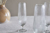 Four Otuba Champagne Flutes by nkuku, crafted from clear recycled glass, sit atop a light textured tablecloth. In the background, a tan and white vase with dried grasses adds elegance to the glassware display.