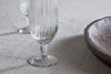 A close-up of two Otuba Champagne Flutes - Clear (Set of 4) by nkuku, made from ribbed recycled glass, displayed on a textured beige surface beside a round wooden bowl.