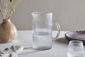 An Otuba Jug - Clear by nkuku, half-filled with water, rests on a light-colored textured tablecloth beside a matching glass, a beige ceramic vase with dried flowers, a napkin, and a wooden bowl.