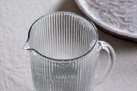 The Otuba Jug - Clear by nkuku, made from recycled glass with a vertical ribbed texture, rests elegantly on a light surface. In the background, a subtly patterned white plate complements its timeless design.