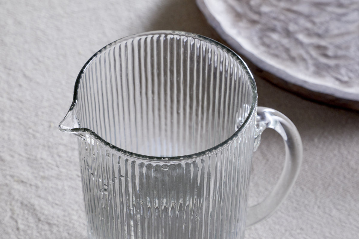 The Otuba Jug - Clear by nkuku, made from recycled glass with a vertical ribbed texture, rests elegantly on a light surface. In the background, a subtly patterned white plate complements its timeless design.