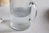 A close-up of the Otuba Jug - Clear by nkuku, featuring a ribbed design and crafted from recycled glass, half-filled with water and set on a textured light-colored tablecloth.