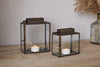 Two nkuku Obi Mini Table Lanterns in antique brass, each with a metal frame and glass door holding a lit tealight, sit on a wooden surface. A vase of dried pampas grass in the background adds to the candlelit ambiance.