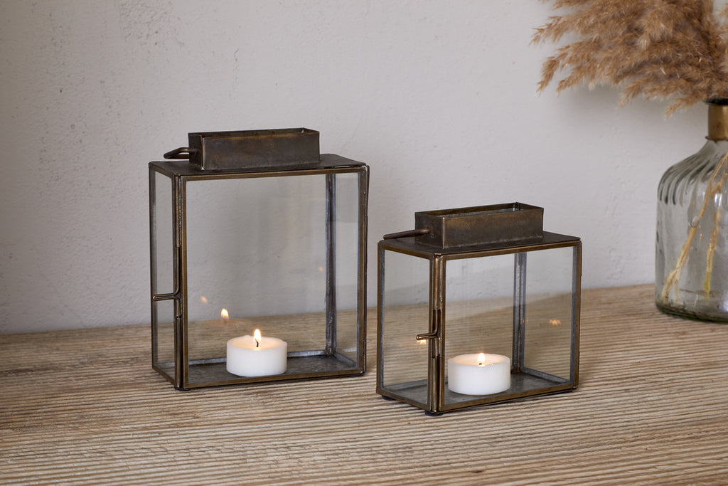 Two nkuku Obi Mini Table Lanterns in antique brass, each with a metal frame and glass door holding a lit tealight, sit on a wooden surface. A vase of dried pampas grass in the background adds to the candlelit ambiance.