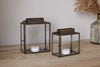 Two nkuku Obi Mini Table Lanterns in Antique Brass with metal frames and glass doors sit on a wooden surface, each holding a white tealight candle. A vase of dried pampas grass adds warmth to the scene.