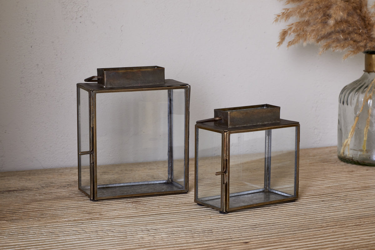 Two nkuku Obi Mini Table Lanterns in antique brass sit on a wooden surface, ready for candlelight. Behind them, a vase with dried pampas grass rests against a light-colored wall.