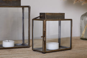 The nkuku Obi Mini Table Lantern in Antique Brass rests on a wooden surface, displaying a white tealight candle inside its glass door. Another similar lantern sits nearby, both ready to cast ambient candlelight.