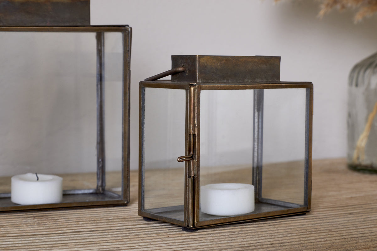 The nkuku Obi Mini Table Lantern in Antique Brass rests on a wooden surface, displaying a white tealight candle inside its glass door. Another similar lantern sits nearby, both ready to cast ambient candlelight.