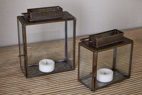 The Obi Mini Table Lantern in Antique Brass by nkuku features a glass door and holds a white tealight candle, casting gentle candlelight on a striped wooden surface against a plain wall.