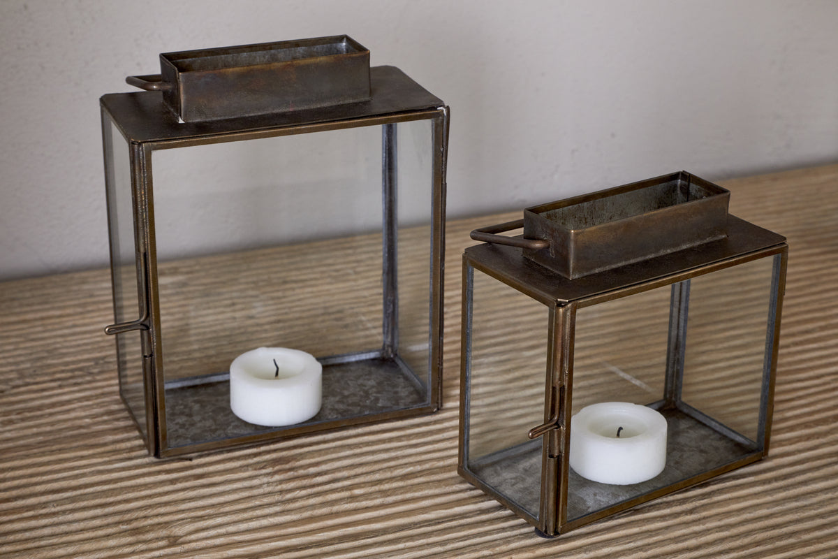 The Obi Mini Table Lantern in Antique Brass by nkuku features a glass door and holds a white tealight candle, casting gentle candlelight on a striped wooden surface against a plain wall.