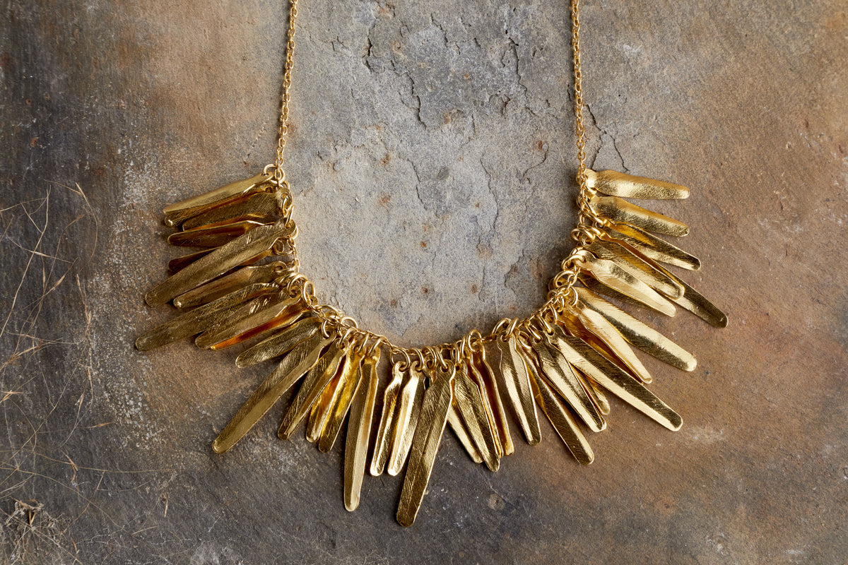 The nkuku Omuti Necklace - Gold features multiple elongated, flat, textured pendants in a bold gold finish, hanging closely together on a chain and displayed on a stone surface.