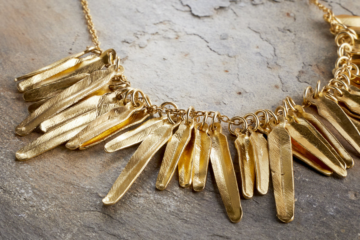 The Omuti Necklace - Gold by nkuku features multiple elongated, textured gold pendants on a chain, shown in close-up on a stone surface.