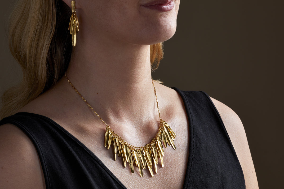 A woman in a black sleeveless top models the nkuku Omuti Necklace - Gold, paired with matching gold earrings featuring bold, elongated shapes, set against a dark background.
