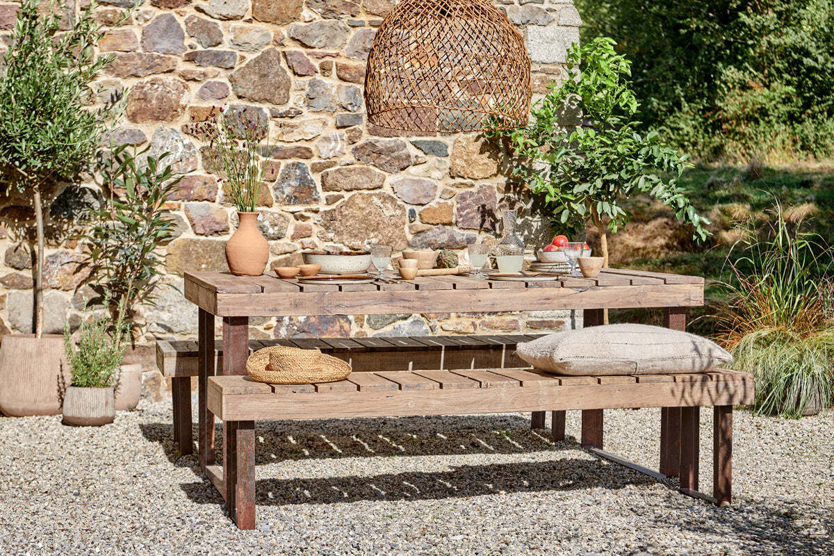 The nkuku Oso Wooden Dining Table with benches stands on gravel by a stone wall, set with ceramic dishes and vases. Bench cushions, green plants, and a woven pendant lamp complete the cozy outdoor scene in natural light.