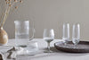 A round tray holds a ribbed glass pitcher of water, two tall ribbed glasses, and Otuba Tumblers – Clear (Set of 4) by nkuku. A tan vase with dried flowers completes this elegant tableware setting on a textured tablecloth.