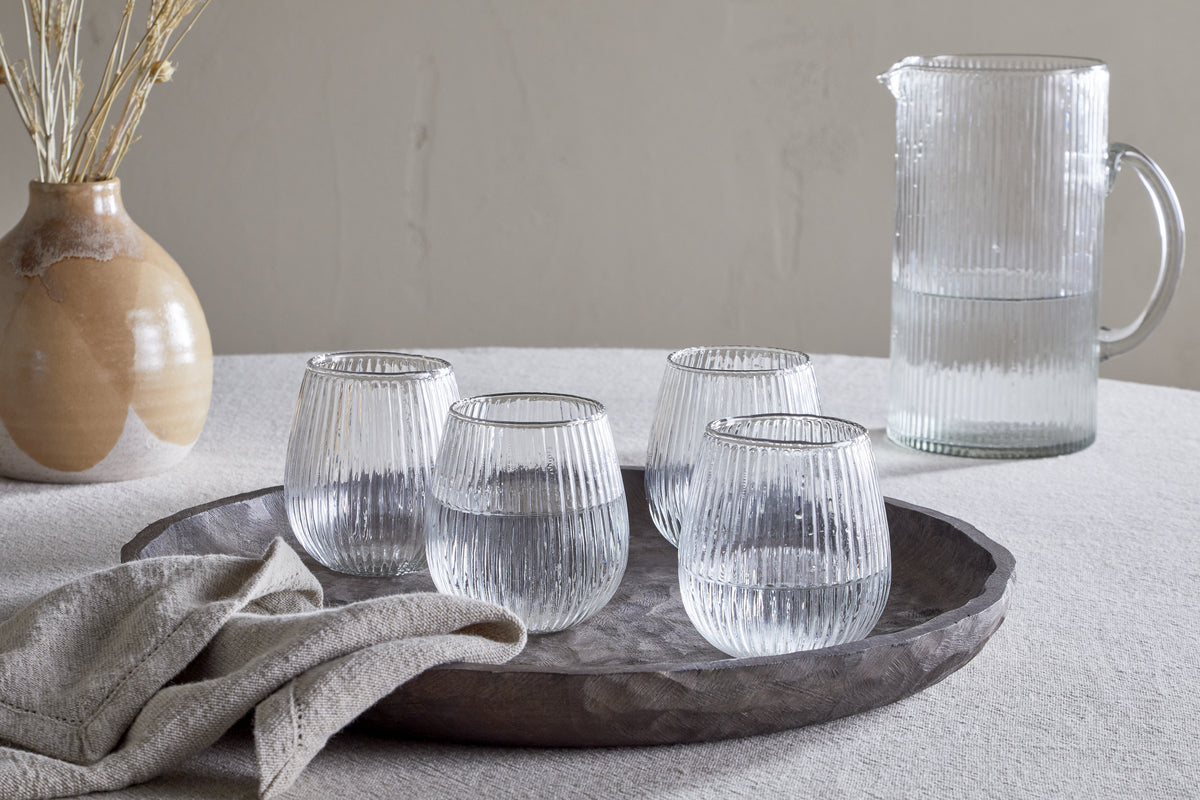 A textured tablecloth holds a round tray with nkuku’s Otuba Tumbler - Clear (Set of 4) made from recycled ribbed glass, next to a folded beige napkin. In the background sit a matching ribbed glass pitcher and a ceramic vase with dried stems.