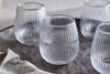 A set of four nkuku Otuba Tumblers in clear, each featuring a ribbed texture, are arranged with water on a round wooden tray. A beige textured cloth accents the scene, complementing this elegant tableware display.