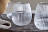 Four Otuba Tumblers - Clear by nkuku, each crafted from recycled glass with a vertical ribbed design, are filled with water and arranged on a dark tray, accented by a beige cloth in the foreground—ideal for stylish table settings.
