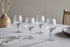 A set of four Otuba Wine Glasses - Clear by nkuku, crafted from recycled glass, are displayed on a light tablecloth with a ceramic vase of dried flowers, a cloth napkin, wooden bowl, and woven chair in the background.