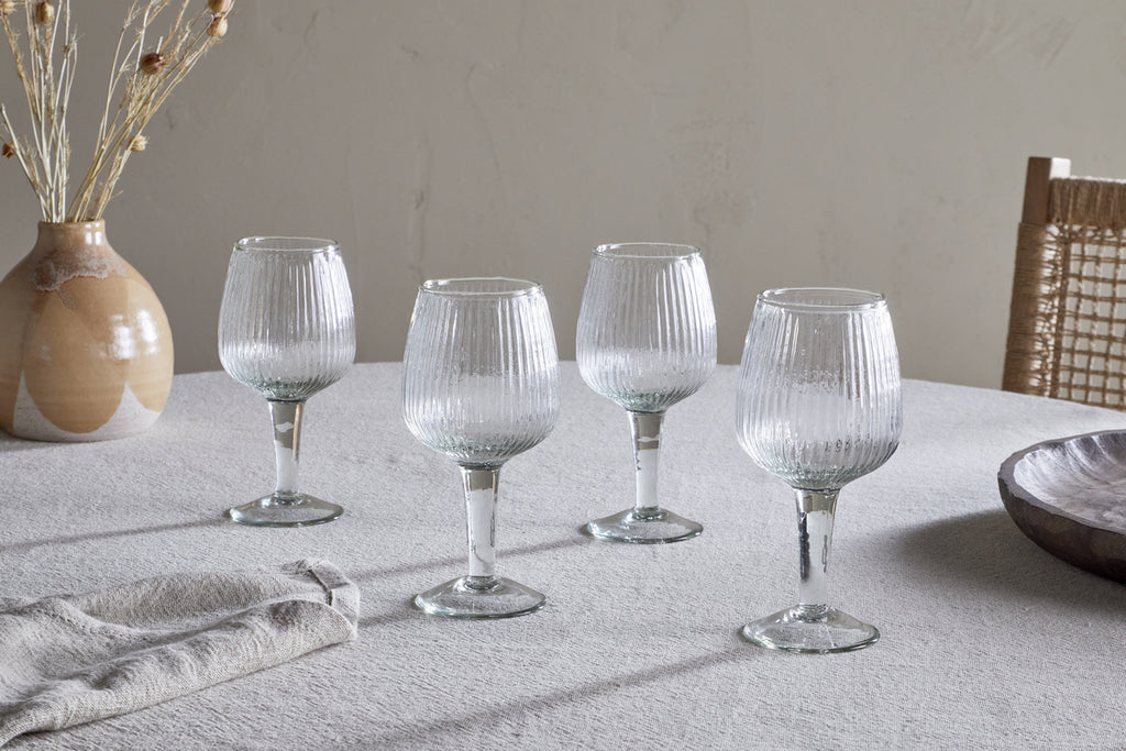 A set of four Otuba Wine Glasses - Clear by nkuku, crafted from recycled glass, are displayed on a light tablecloth with a ceramic vase of dried flowers, a cloth napkin, wooden bowl, and woven chair in the background.