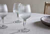Four elegant, ribbed Otuba Wine Glasses by nkuku sit on a light textured tablecloth, with a wooden bowl and woven chair in the background for a serene, minimal look.