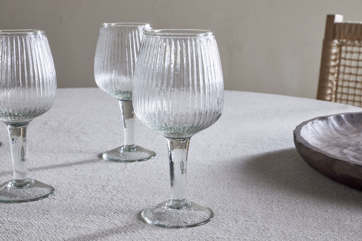 Four elegant, ribbed Otuba Wine Glasses by nkuku sit on a light textured tablecloth, with a wooden bowl and woven chair in the background for a serene, minimal look.