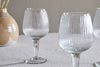 Two Otuba Wine Glasses by nkuku (Clear, Set of 4) sit on a light textured tablecloth, with a tan and white ceramic vase in the background. Another wine glass is partially visible on the right side.