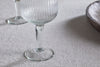 A close-up of the elegant Otuba Wine Glass - Clear (Set of 4) by nkuku, featuring ribbed recycled glass on a textured, light tablecloth, with another glass and a wooden bowl in the background.