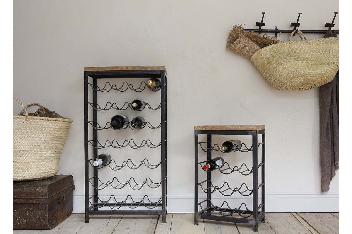 Two nkuku Obra Industrial Wine Racks—crafted from mango wood and iron in a small size—stand on a wooden floor against a light wall, holding several bottles; wicker baskets and a brown blanket hang above vintage chests and extra storage.