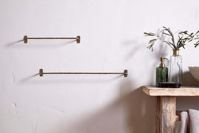 A minimalist bathroom features two nkuku Olani Bathroom Hanging Rails in antique brass on a white wall. To the right, a rustic wood shelf holds a glass bottle with olive branches, a green soap dispenser, and neatly folded towels.