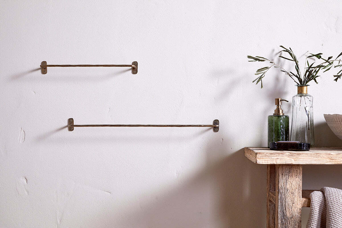 A minimalist bathroom features two nkuku Olani Bathroom Hanging Rails in antique brass on a white wall. To the right, a rustic wood shelf holds a glass bottle with olive branches, a green soap dispenser, and neatly folded towels.
