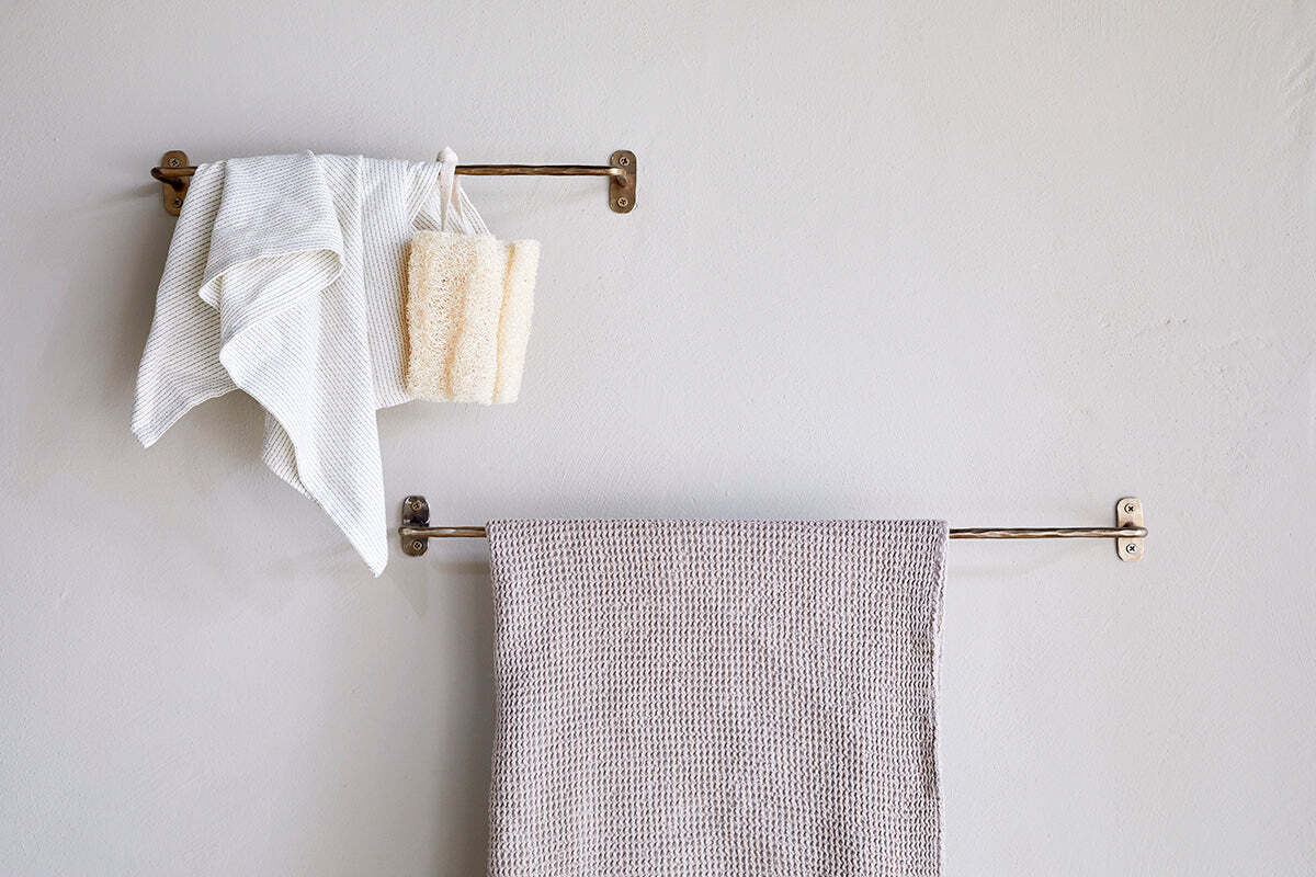 Two nkuku Olani Bathroom Hanging Rail racks in antique brass are mounted on a light bathroom wall. The top rack holds a white towel, washcloth, and beige bath sponge; the lower rack displays a textured beige towel. The wall is unadorned.