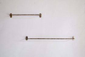 Two nkuku Olani Bathroom Hanging Rails in antique brass are mounted on a light bathroom wall; the upper rail is shorter and to the left, while the lower, longer rail is offset to the right, both fixed with visible screws.