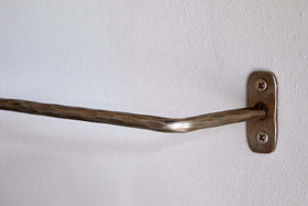 A close-up of an nkuku Olani Bathroom Hanging Rail in antique brass with a hammered texture, mounted on a white wall with a rectangular bracket and two visible screws; the bar is slightly bent, and the wall surface is smooth.