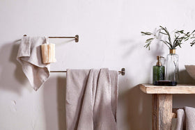 A minimal bathroom scene featuring beige towels on the nkuku Olani Bathroom Hanging Rail in Antique Brass, a loofah, and a rustic wooden shelf with a green glass soap dispenser, olive branches in a vase, and a bowl against a light wall.